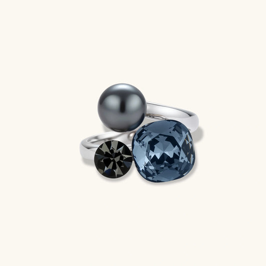 Adjustable ring with blue cushion-cut zirconia, gray pearl, and dark crystal on platinum-plated copper band against beige background.